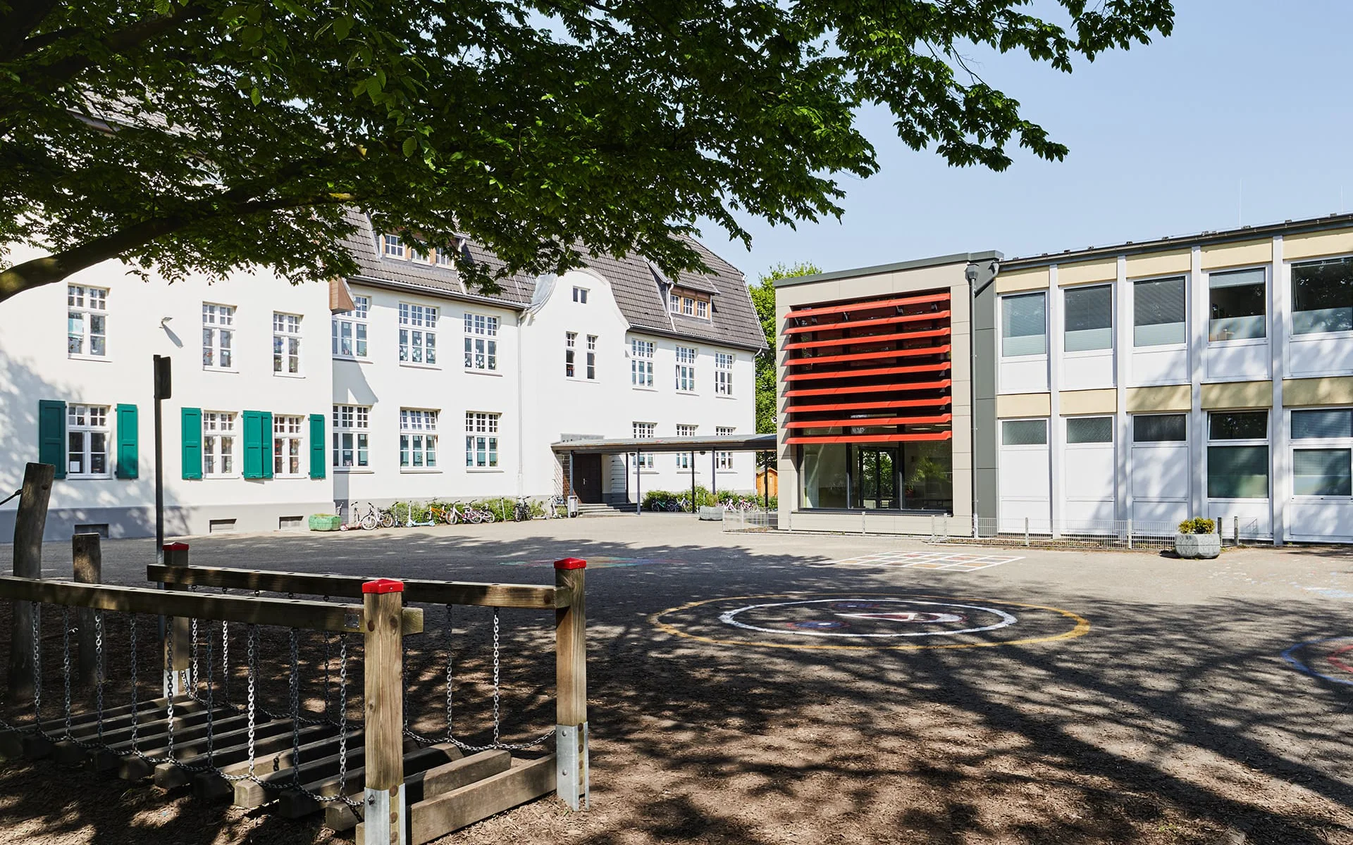 Schule mit roten Lamellen an Fensterfront und Spielplatz vorne links